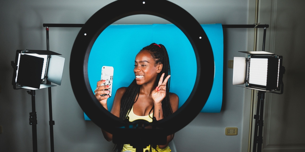 In a studio, a ring light frames a young Black female influencer who is smiling while looking into the mobile phone that she is holding.